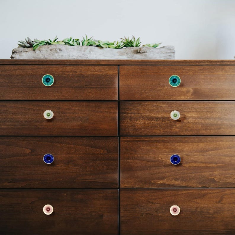 May include: A dark brown wooden dresser with six drawers, each adorned with a unique, colorful knob. The knobs feature shades of teal, green, blue, and pink, adding a pop of color to the furniture. A rectangular planter with greenery sits on top.