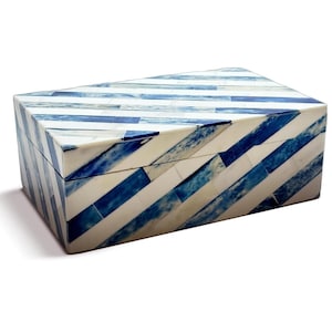 May include: Rectangular box with a slanted lid, featuring a blue and white striped pattern. The stripes are arranged diagonally across the surface, creating a geometric design. The box appears to be made of a hard material.