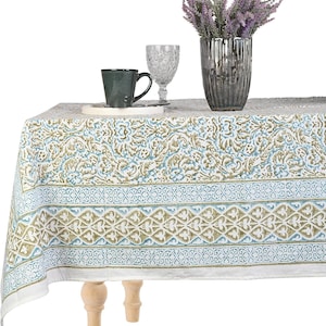 May include: A white tablecloth with a blue and olive green floral pattern, set with a dark green mug, a clear glass, and a silver vase with purple flowers. The table has light wood legs.