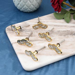 May include: A collection of decorative cabinet knobs. Each knob features a gold-colored metal base with a stylized floral design, inlaid with a pearlescent material. The knobs are arranged on a marble-patterned surface, suggesting a home decor theme.