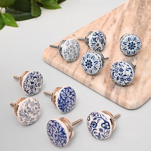 May include: A collection of nine ceramic cabinet knobs with floral designs in white and blue. The knobs have metal stems and are arranged on a marble-like surface. The designs include various floral and leaf patterns.