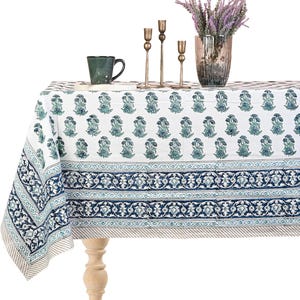 May include: A white tablecloth with a blue floral pattern and a blue border. The table is set with a green mug, gold candlesticks, and a vase of purple flowers. The tablecloth is draped over a wooden table.