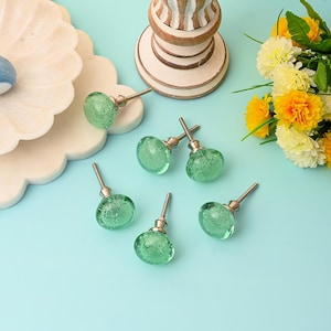 Peut inclure: Six boutons de meuble en verre vert avec des tiges argentées sont disposés sur une surface bleu clair. Les boutons ont une texture à bulles et sont ronds. Un plateau décoratif blanc et un support en bois sont également dans l'image, avec des fleurs jaunes et blanches.