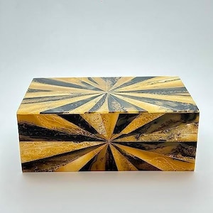 May include: A rectangular box featuring a sunburst pattern of alternating black and yellow segments. The surface displays a marbled texture throughout the design, with the pattern radiating from a central point on both the top and front panels.