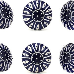 Peut inclure: Six boutons de meuble en céramique bleu marine et blanc. Chaque bouton présente un motif circulaire avec un centre bleu foncé et un motif de formes en goutte d'eau blanches rayonnant vers l'extérieur. Les boutons ont des tiges métalliques dorées.