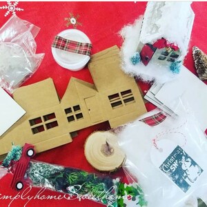 May include: A collection of Christmas craft supplies on a red tablecloth. Includes cardboard house cutouts, white envelopes, faux snow, a miniature red truck, and a small bottle brush tree. A tag reads "Christmas Snow".