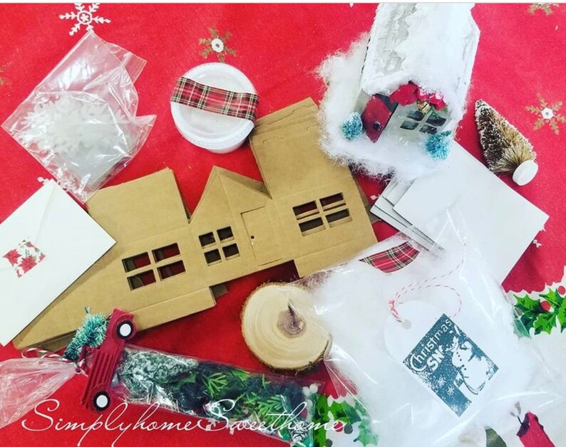 May include: A collection of Christmas craft supplies on a red tablecloth. Includes cardboard house cutouts, white envelopes, faux snow, a miniature red truck, and a small bottle brush tree. A tag reads "Christmas Snow".