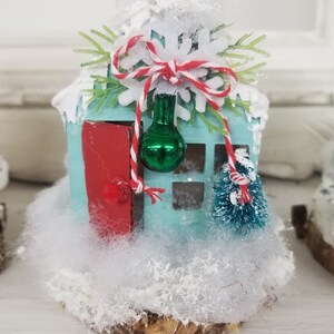 May include: A miniature, light blue house with a red door and a small green Christmas tree. The house is decorated with white snow, a red and white string, and a green ornament. The house sits on a wooden base, creating a festive holiday decoration.