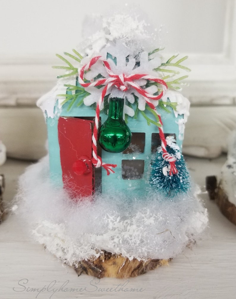 May include: A miniature, light blue house with a red door and a small green Christmas tree. The house is decorated with white snow, a red and white string, and a green ornament. The house sits on a wooden base, creating a festive holiday decoration.