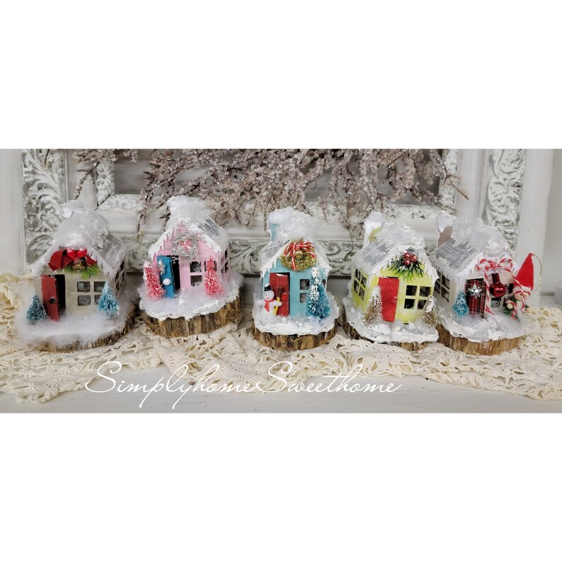 May include: Five miniature Christmas village houses with snow-covered roofs and bases. Each house has a different color scheme, including red, pink, blue, yellow, and white, with decorative wreaths and miniature trees. The houses are set on wooden bases.
