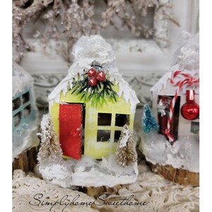 May include: A collection of miniature Christmas village houses. The houses are painted in various colors, including yellow and white, and are covered in artificial snow. Each house has a red door and is decorated with small Christmas trees and berries. The text "Simplyhome Sweet home" is at the bottom.