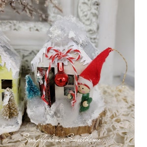 May include: A miniature white Christmas house ornament with a red and white striped bow, a red ornament, and a small elf figure wearing a red hat and green coat. The house is decorated with faux snow and a small blue Christmas tree. The base is a wooden slice.