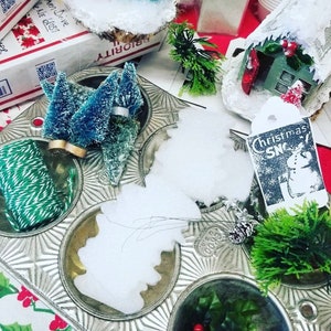May include: A vintage muffin tin holds miniature Christmas decorations, including bottle brush trees, faux snow, and holly. The scene includes a tag that reads "Christmas Snow" and a small house. The background includes Priority Mail boxes.