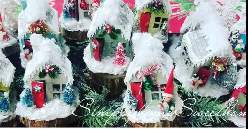 May include: A collection of miniature Christmas houses covered in faux snow. Each house has a red door, some with small Christmas trees and decorative accents. The houses are set on wooden bases and surrounded by greenery.