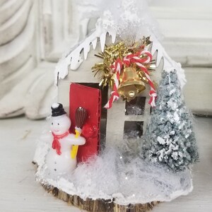 May include: A miniature Christmas house with a red door, a snowman, and a small Christmas tree. The house is decorated with white frosting and a gold bell. The base is a wooden slice, and the scene is covered in faux snow.