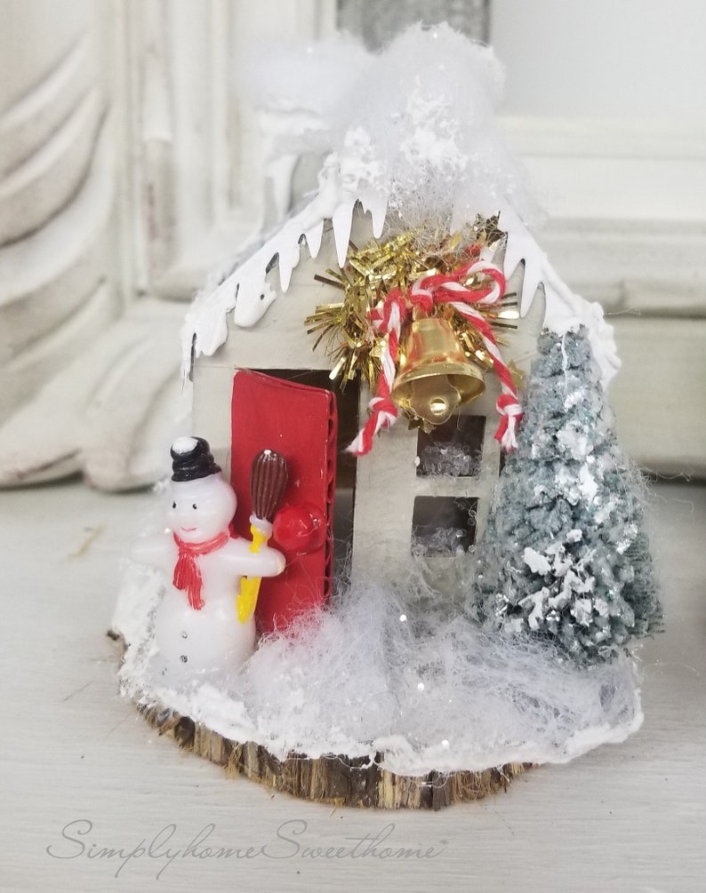 May include: A miniature Christmas house with a red door, a snowman, and a small Christmas tree. The house is decorated with white frosting and a gold bell. The base is a wooden slice, and the scene is covered in faux snow.