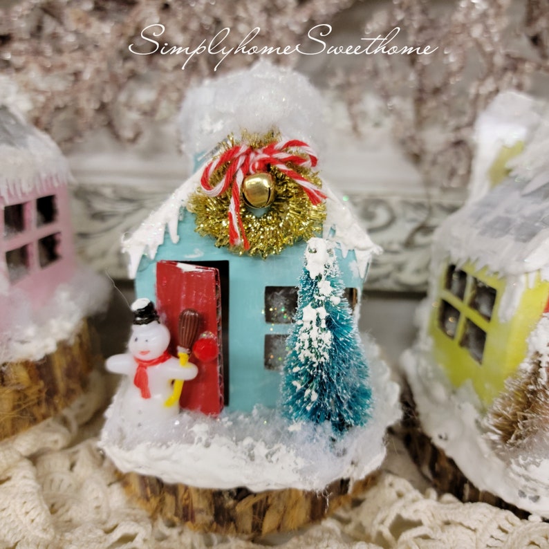 May include: A miniature, light blue Christmas house with a red door, a gold tinsel wreath, and a small snowman figurine. The house is covered in snow and sits on a wooden base. The text "Simplyhome Sweet home" is at the top.
