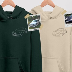 May include: Two hooded sweatshirts, one dark green and one beige, displayed on wooden hangers. Each sweatshirt has a line drawing of a car on the front. Small photos of cars are taped to the sweatshirts. The text "YOUR PHOTO" is visible on the photos.