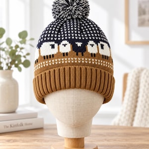 May include: A knit beanie featuring a dark blue top section with a white sheep pattern, a brown middle band with a white geometric design, and a solid brown ribbed cuff. The hat is topped with a dark blue and white pom-pom.