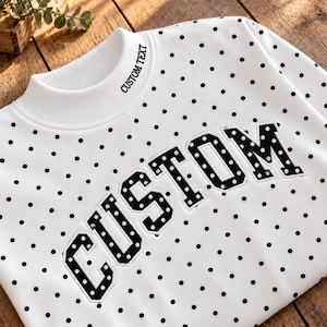 May include: A white long-sleeve sweatshirt featuring a black polka dot pattern throughout. The front displays the word CUSTOM in large black block letters with white dots, and the mock neck collar includes the text CUSTOM TEXT.