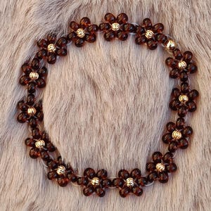 May include: A necklace featuring a floral design. The necklace is composed of dark brown, flower-shaped beads with gold-colored centers. The beads are strung together to form a circular shape.