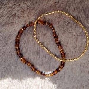 May include: Two beaded bracelets. One bracelet features small, gold-colored beads. The other bracelet has a mix of amber and brown-colored beads. The bracelets are displayed on a textured, light brown surface.