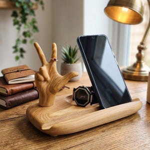May include: A light brown wooden desk organizer featuring a hand sculpture making a peace sign, a slot for a smartphone, and a recessed area for a wristwatch. The hand sculpture is adorned with a small ring featuring a red stone.