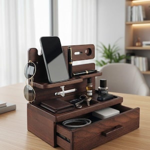 May include: A dark brown wooden desk organiser with multiple compartments and a drawer. It holds a smartphone, pens, wallet, keys, and other small items. Sunglasses hang on the side. The organiser is on a light brown wooden surface.