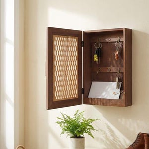 May include: A wooden key holder with a hinged door featuring a decorative, openwork design. The interior has hooks for keys and a slot for mail. The key holder is mounted on a beige wall.