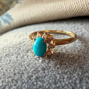 May include: A gold ring featuring an oval, bright turquoise gemstone at the center, surrounded by smaller, clear stones. The ring is set against a soft, gray background.