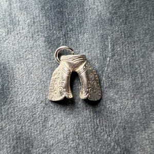 Op de afbeelding: Een zilverkleurige bedel in de vorm van een broek. De bedel heeft ingewikkelde bloemen- en stippenpatronen op de benen. Er is een kleine ring aan de bovenkant bevestigd om op te hangen. De bedel is waarschijnlijk van metaal.