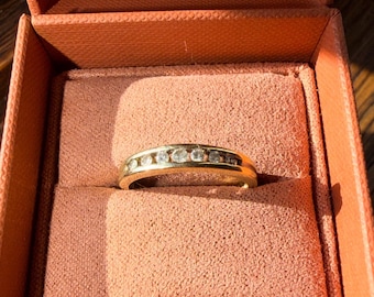 May include: A gold band ring with seven clear, round-cut gemstones set in a channel. The ring is displayed inside a peach-colored jewelry box with a velvet lining. The box is open, revealing the ring.