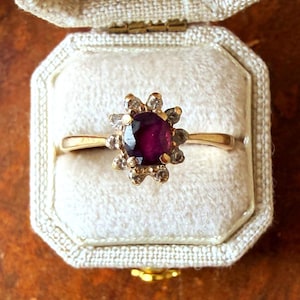 May include: A gold ring featuring a central oval-cut deep red gemstone surrounded by a halo of ten small, clear, round-cut stones. The ring has a polished gold band and is presented in a cream-colored, textured, octagonal jewelry box.
