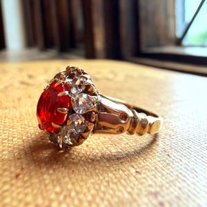 Könnte beinhalten: Ein goldener Ring mit einem großen, oval geschliffenen roten Edelstein, umgeben von kleineren, klaren Steinen. Der Ring hat eine verzierte, detaillierte Fassung und einen polierten Ring. Der Ring liegt auf einer strukturierten, hellbraunen Oberfläche.