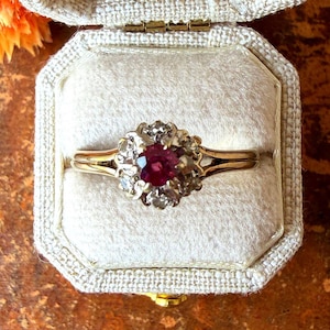 May include: A gold ring featuring a central round-cut red ruby surrounded by a cluster of small, clear, round-cut diamonds. The ring has a polished gold band that tapers toward the center, showcasing a classic floral-inspired cluster setting.