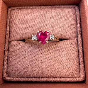 Puede incluir: Un anillo de oro con una piedra preciosa roja vibrante en forma de corazón en el centro, flanqueada por dos piedras claras más pequeñas, de corte cuadrado. El anillo se presenta en una caja de joyería rosa.