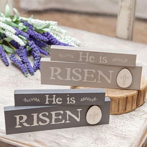 May include: Two gray wooden blocks with the words "He is RISEN" in white lettering. Each block has a decorative egg with a cross. A bouquet of purple and white flowers is in the background. Easter decor.