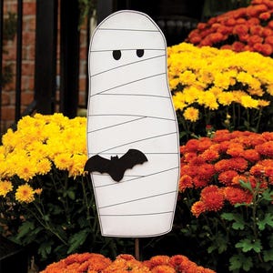 May include: A white mummy-shaped Halloween yard decoration with black eyes and a black bat attached. The mummy is wrapped in white bandages with black lines. The decoration is placed in a garden with orange and yellow flowers.