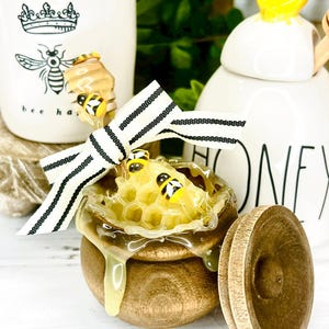 May include: A wooden honey pot with a honeycomb design and dripping honey. The pot is adorned with a black and white striped bow and decorative bees. A white ceramic jar with a bee graphic is in the background.