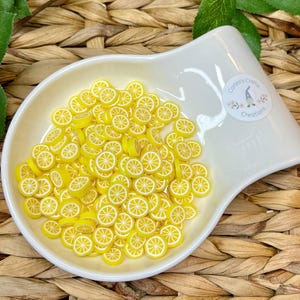 May include: A white ceramic dish filled with yellow lemon slices. The lemon slices have white details and are arranged in a decorative pattern. The dish has a curved shape and a small logo that says "Carlee's Crafty Creations".