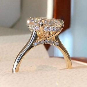 May include: A gold engagement ring with a large, oval-cut diamond. The ring has a halo of smaller diamonds around the centre stone and along the band. The ring is set in a gold band and is displayed in a ring box.