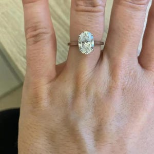 May include: A rose gold ring featuring a large, oval-cut diamond. The diamond is held in place by four prongs and reflects light. The ring is worn on a finger, highlighting the sophisticated design and the diamond's sparkle.