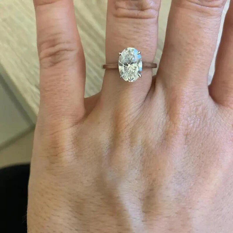 May include: A rose gold ring featuring a large, oval-cut diamond. The diamond is held in place by four prongs and reflects light. The ring is worn on a finger, highlighting the sophisticated design and the diamond's sparkle.