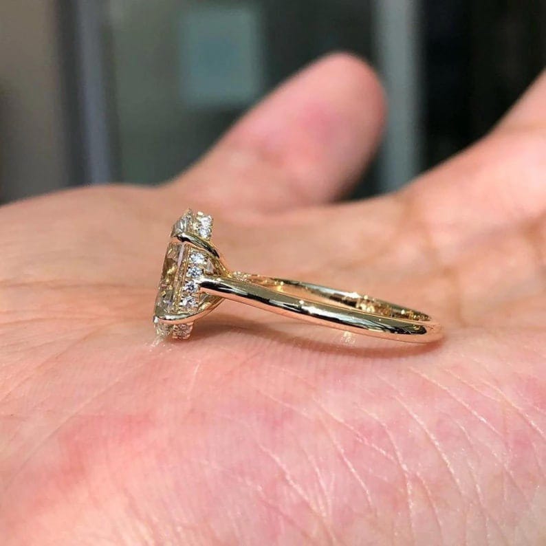 May include: A gold ring featuring a large, light-coloured central stone and smaller accent stones. The ring's band is thin and polished, with intricate setting details. The ring is held in a hand, highlighting its design and craftsmanship.