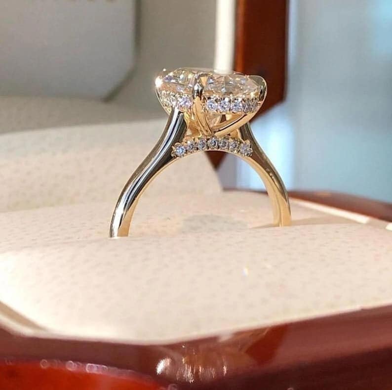 May include: A gold engagement ring with a large, oval-cut diamond. The ring has a halo of smaller diamonds around the centre stone and along the band. The ring is set in a gold band and is displayed in a ring box.