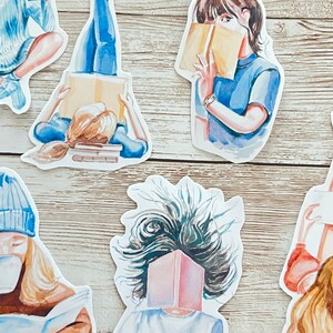 May include: A set of five watercolor stickers featuring women reading books. The stickers are on a light wood background. The women are depicted in various poses, some with their hair flowing in the wind, others with their hair pulled back. The books are all different colors and sizes.