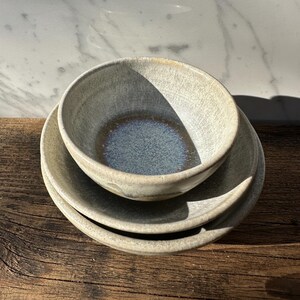 Può includere: Un set di tre ciotole e piatti in ceramica. Gli articoli sono di un colore grigio-verde tenue con una texture maculata. L'interno della ciotola superiore ha una glassa blu scuro. Gli articoli sono disposti su una superficie di legno invecchiato.