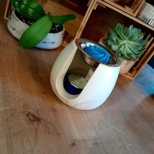 以下が含まれることがあります： 銀色のトップと青いバラの形をしたワックスメルトが付いた、白い卵型のエッセンシャルオイルディフューザー。ディフューザーの中には小さな青いキャンドルがあります。ディフューザーは木製の表面にあり、背景には鉢植えの植物があります。