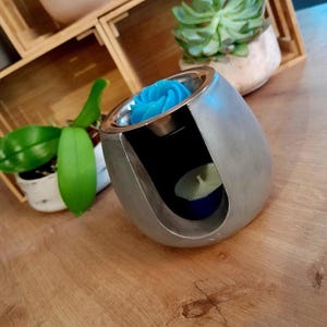 May include: A silver-coloured essential oil diffuser with a teal rose-shaped wax melt. The diffuser has a metallic rim and a tealight candle inside. The design is modern and sleek, suitable for aromatherapy.