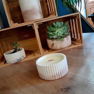 Peut inclure: Une bougie blanche et beige avec un design côtelé et une mèche en bois est posée sur une surface en bois. La bougie est à côté de petites plantes succulentes en pot et de caisses en bois décoratives. La bougie est beige clair.